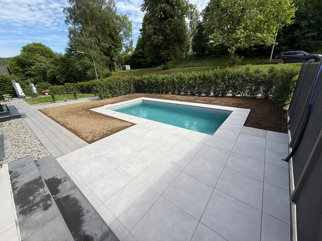 Großformatige helle Plattenterrasse rund um ein Schwimmbecken, dahinter Hecke und Rasen