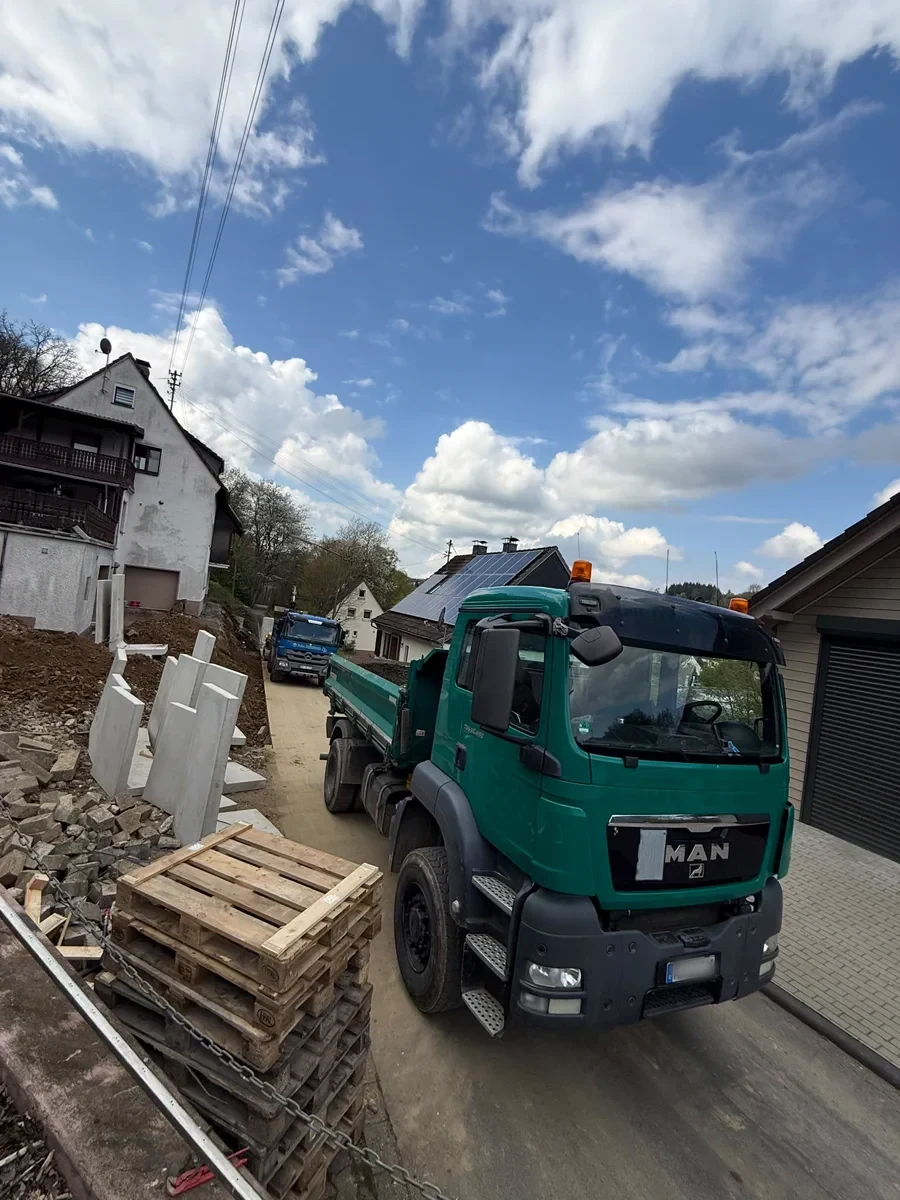 Grüner Dreiseitenkipper des Betriebs auf einer Hausbaustelle, daneben Paletten und Baumaterial