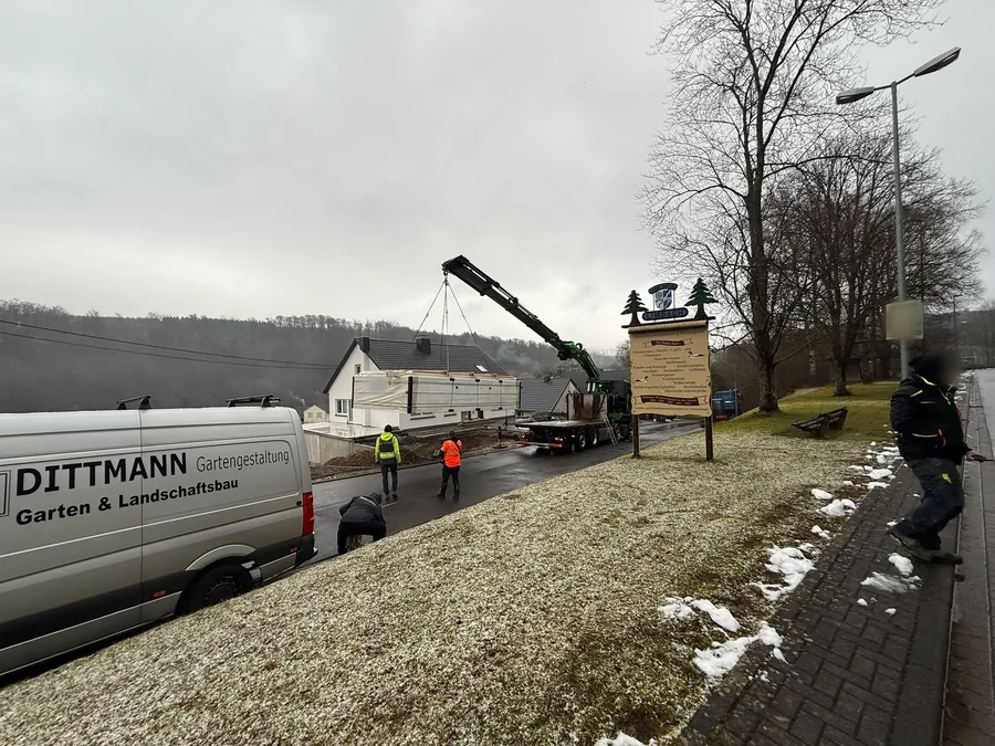 Firmentransporter an der Straße, dahinter hebt ein Ladekran ein Poolbecken auf ein Grundstück