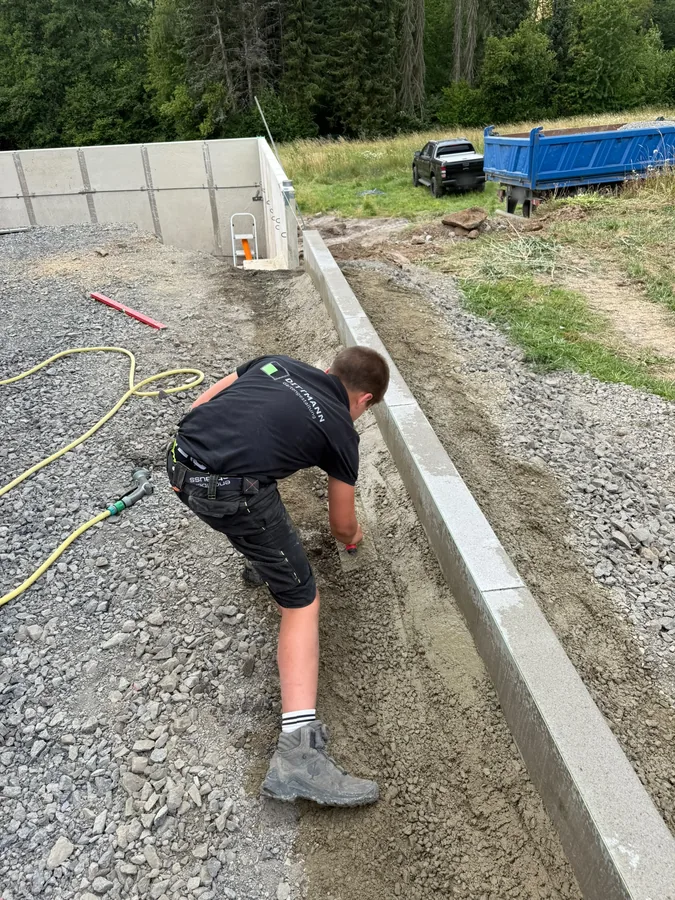 Randsteinreihe wird in frisches Betonbett gesetzt, entlang einer Schotterfläche vor einer Betonwand