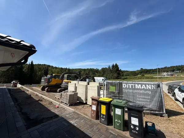 Baustelle vor einem Neubau mit zwei Kettenbaggern, gestapelten Betonwinkelsteinen und Pflasterpaletten