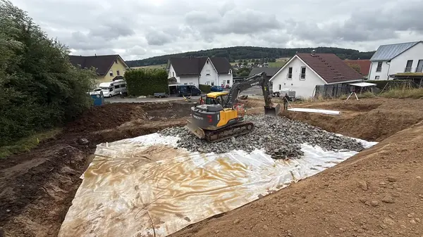 Kettenbagger verteilt Schotter auf einem ausgelegten Geotextil in einer Baugrube am Ortsrand