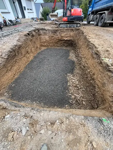 Ausgehobene Baugrube mit eingebrachter Schottertragschicht, oben am Rand ein Minibagger und ein Kipper