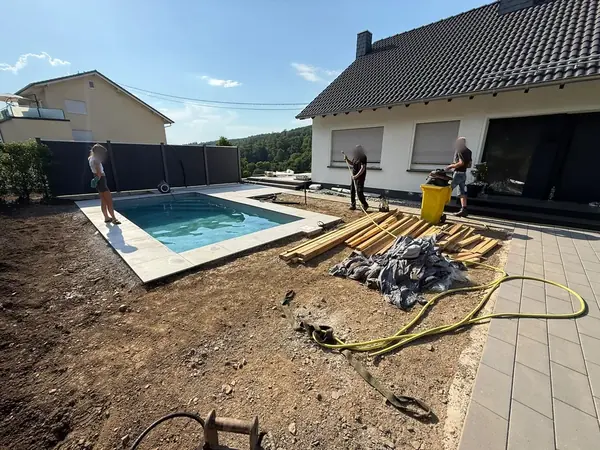 Mit Wasser gefüllter Pool mit umlaufender Steinkante neben einem Wohnhaus, das Umfeld ist noch Rohboden