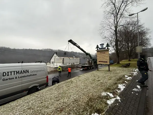 Firmentransporter an der Straße, dahinter hebt ein Ladekran ein Poolbecken auf ein Grundstück
