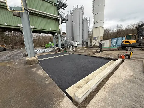 Frisch eingebaute Asphaltfläche mit Betonrinne, rechts ein Kettenbagger
