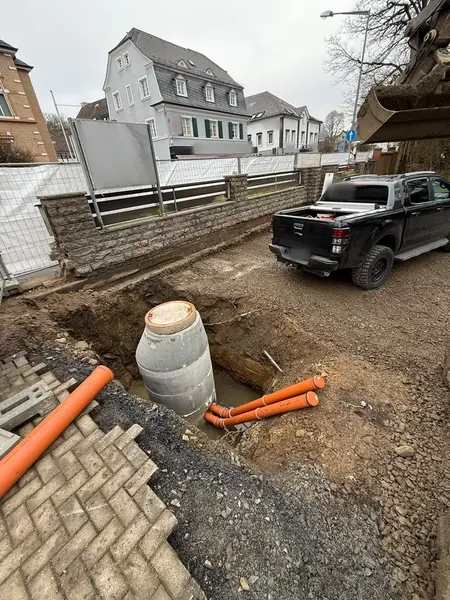 Betonschacht und orangefarbene KG-Rohre in einer offenen Baugrube, daneben der Pick-up des Betriebs